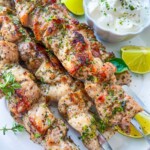 Grilled pork skewers garnished with herbs on a white plate, served with fresh lime wedges and a bowl of creamy white dipping sauce topped with chopped herbs.