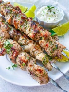 Grilled chicken and salmon skewers seasoned with herbs, served on a white plate with lime wedges and a bowl of creamy dipping sauce, garnished with fresh herbs.