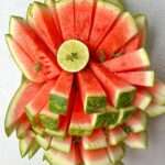 Sliced watermelon arranged in a fan shape on a light surface, topped with a round lime slice and garnished with small green herbs and seasoning, resembles refreshing Watermelon Popsicles perfect for summer.