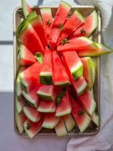 A tray filled with neatly arranged slices of fresh watermelon, resembling watermelon popsicles and garnished with small green herbs, sits on a light surface with sunlight casting playful shadows.
