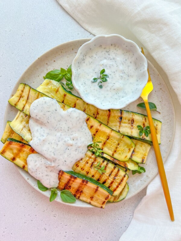 Grilled zucchini slices arranged on a plate with a creamy herb sauce drizzled on top and served in a small bowl, garnished with fresh herbs and a gold spoon on the side.