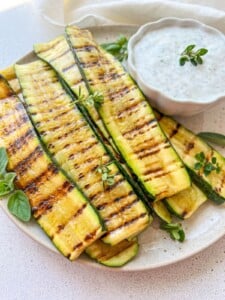 A plate of grilled zucchini slices garnished with fresh herbs, served with a bowl of creamy white dipping sauce topped with sprigs of herbs.