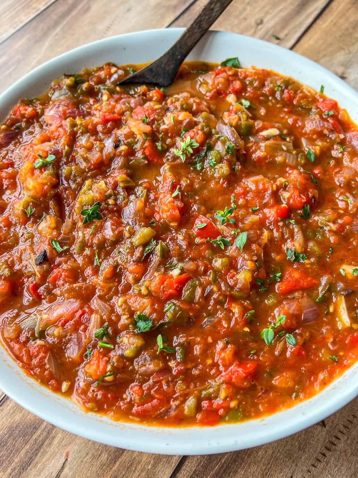 A close-up of a bowl filled with chunky, homemade roasted vegetable salsa, featuring visible pieces of tomato, pepper, onion, and herbs, with a spoon resting inside the bowl on a wooden table.