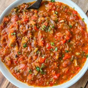 A white bowl filled with chunky, red and green salsa garnished with chopped herbs sits on a wooden surface, with a dark spoon resting in the bowl.