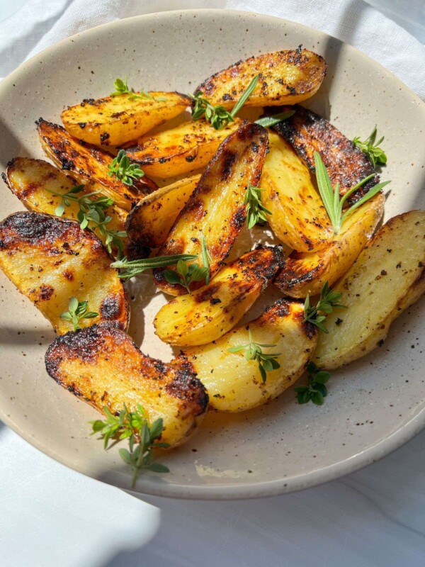 Golden roasted potato wedges, inspired by Grilled Greek Potatoes and garnished with fresh herbs, served in a speckled beige bowl under bright natural light.