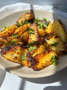 A beige bowl filled with golden, Grilled Greek Potatoes garnished with fresh green herbs sits on a white surface in natural sunlight.