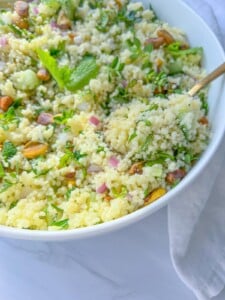 A close-up of a bowl of couscous salad with chopped red onions, cucumber, fresh herbs, and pistachios, inspired by Cranberry Quinoa Salad, with a gold fork resting in the bowl on a white surface.