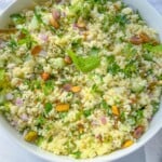 A white bowl filled with fresh Jennifer Aniston Couscous Salad, featuring chopped cucumber, red onion, herbs, and garnished with pistachios and mint leaves. The bowl is set on a white surface.