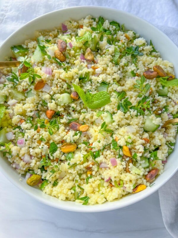 A white bowl filled with fresh Jennifer Aniston Couscous Salad, featuring chopped cucumber, red onion, herbs, and garnished with pistachios and mint leaves. The bowl is set on a white surface.