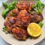 A plate of Lemon Pepper Chicken thighs garnished with fresh basil and herbs, served with a halved lemon seasoned with black pepper on the side.