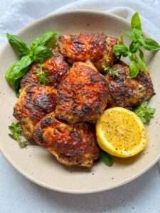 A plate of Lemon Pepper Chicken thighs garnished with fresh basil and herbs, served with a halved lemon seasoned with black pepper on the side.