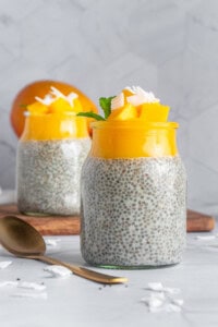 Two jars of chia pudding topped with mango chunks, coconut flakes, and mint leaves are placed on a white surface. A golden spoon lies beside them. A whole mango is seen in the background on a wooden board. The backdrop is a subtle, patterned wall.