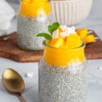 Two glass jars filled with chia pudding topped with mango chunks, coconut flakes, and mint leaves. A gold spoon lies beside them. In the background, a bowl of mangoes sits on a wooden board.