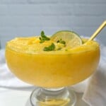 A glass dessert bowl filled with creamy yellow mango frozen yogurt mousse, garnished with a lime slice and fresh green herbs, with a gold spoon resting inside. The background is light and minimal.