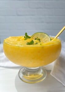 A glass dessert bowl filled with creamy yellow mango frozen yogurt mousse, garnished with a lime slice and fresh green herbs, with a gold spoon resting inside. The background is light and minimal.