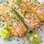 Three grilled corn cobs on skewers, Mexican Street Corn style, coated with creamy sauce, grated cheese, herbs, and chili powder, served on a white plate with lime wedges and sprinkled with chopped parsley.