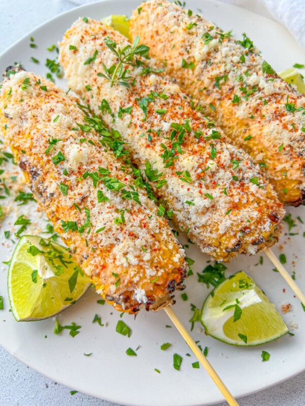 Three grilled corn cobs on skewers, Mexican Street Corn style, coated with creamy sauce, grated cheese, herbs, and chili powder, served on a white plate with lime wedges and sprinkled with chopped parsley.