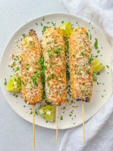 Three Mexican Street Corn (elote) skewers, coated with cheese, spices, and herbs, are served on a white plate with lime wedges and a garnish of chopped parsley. A white napkin rests beside the plate.