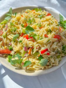 A bowl of Orzo Pasta Salad with chopped red and green bell peppers, red onions, fresh herbs, and a light dressing, garnished with sprigs of oregano on a white background.
