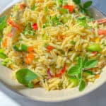 A vibrant Orzo Pasta Salad featuring diced cucumber, tomato, and red onion, garnished with fresh herbs on a white plate. This bright, colorful dish looks as fresh as it tastes.