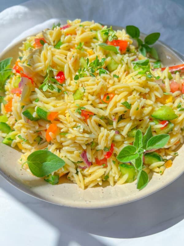 A vibrant Orzo Pasta Salad featuring diced cucumber, tomato, and red onion, garnished with fresh herbs on a white plate. This bright, colorful dish looks as fresh as it tastes.