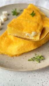 A golden Feta and Potato Omelet with a creamy cheese filling is served on a speckled plate. A fork holds up a bite-sized piece, showing the soft interior. Small herb sprigs and cheese cubes are on the side.