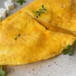 A golden-yellow Feta and Potato Omelet garnished with fresh herbs is served on a speckled plate, with cubes of white cheese and green sprigs on the side. A gold fork rests on the omelet.