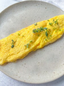 A neatly folded yellow omelet garnished with small green herbs sits on a speckled light gray ceramic plate, accompanied by perfectly cooked Air Fryer Hard Boiled Eggs for a delicious breakfast spread.