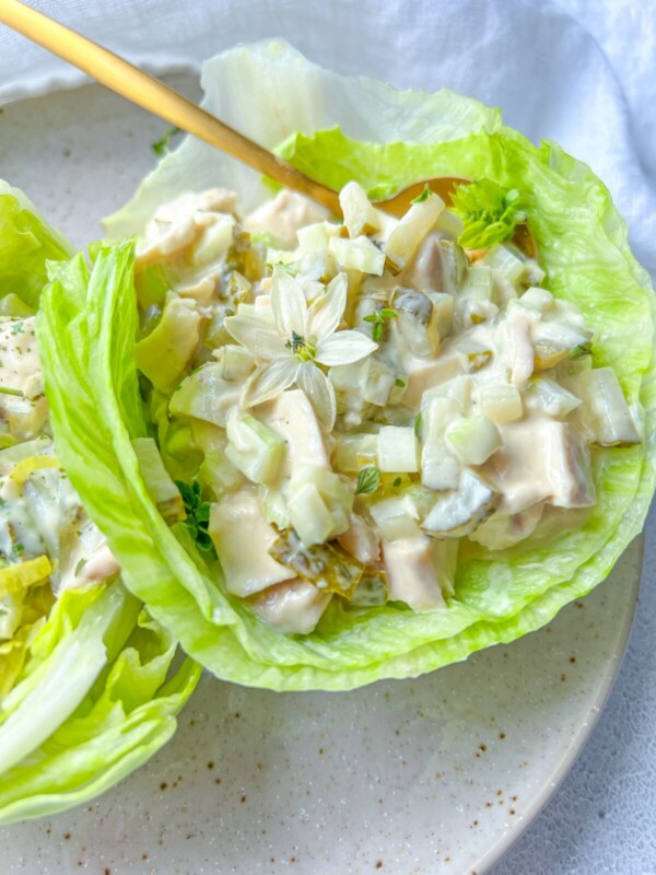 A close-up of Chicken Lettuce Cups featuring creamy chicken salad with diced vegetables, served in fresh lettuce leaves and garnished with a small white edible flower, on a beige plate.