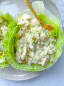 A lettuce leaf filled with creamy chicken salad inspired by Olivier Salad, featuring diced chicken, celery, and pickles, garnished with edible flowers and herbs, on a beige plate with a gold spoon.