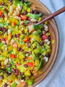 A wooden bowl filled with vibrant Cowboy Caviar, featuring black beans, white beans, corn, bell peppers, celery, fresh herbs, and a wooden spoon on the side.