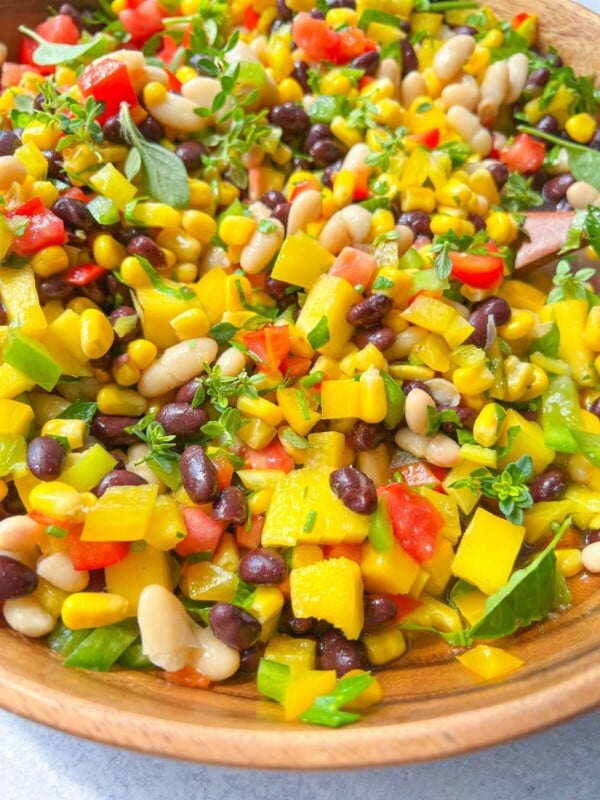 A close-up of Cowboy Caviar in a wooden bowl, featuring black beans, white beans, corn, diced red and yellow bell peppers, tomatoes, and fresh green herbs.