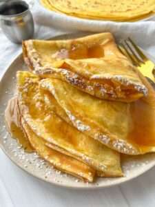 A plate of golden crepes, made from an Easy Crepe Recipe, folded and topped with powdered sugar and syrup, with a gold fork and a small pitcher of syrup beside the plate on a white table.