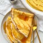 A plate with folded crepes drizzled with syrup and lightly dusted with powdered sugar, set beside a gold fork and knife. Made using an Easy Crepe Recipe, a stack of crepes and a container of syrup rest in the background on a white cloth.