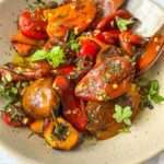 A beige bowl filled with roasted tomatoes, eggplants, and roasted bell peppers garnished with fresh herbs sits on a white surface, with a wooden spoon resting in the bowl.
