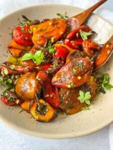 A beige bowl filled with roasted tomatoes, eggplants, and roasted bell peppers garnished with fresh herbs sits on a white surface, with a wooden spoon resting in the bowl.