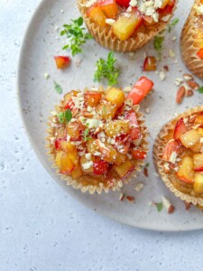 Overhead view of Almond Flour Blueberry Muffins topped with diced fresh peaches and crushed nuts, arranged on a speckled ceramic plate with a few green herb sprigs scattered around.