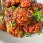 A bowl of baked chicken thighs covered in a chunky, red tomato-based sauce—perfect for an Ajvar Chicken Recipe—garnished with fresh green herbs and served with a gold fork on a speckled plate.