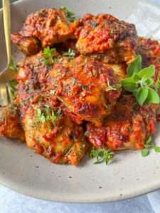 A bowl of baked chicken thighs covered in a chunky, red tomato-based sauce—perfect for an Ajvar Chicken Recipe—garnished with fresh green herbs and served with a gold fork on a speckled plate.