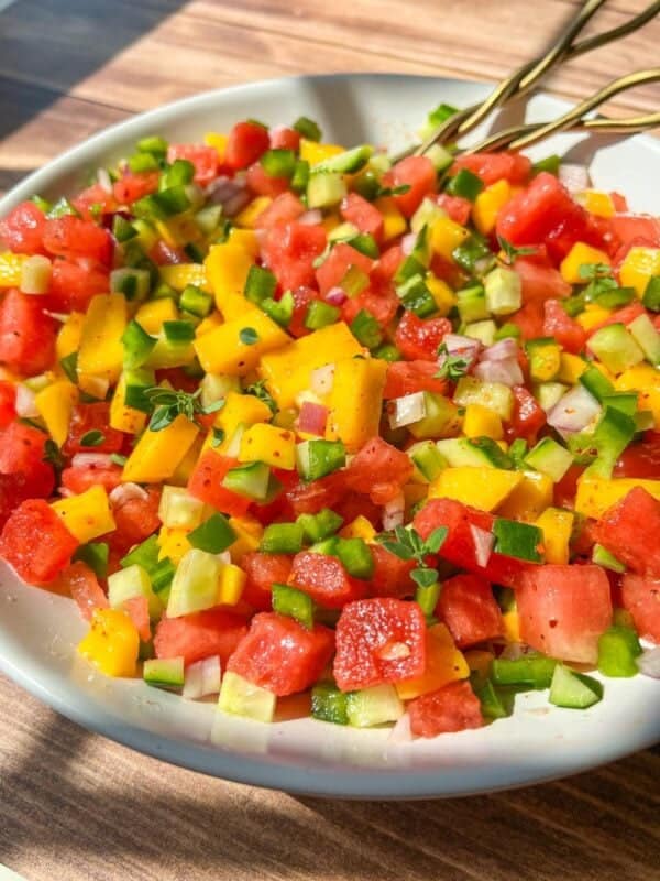 A close-up of a colorful salad with diced watermelon, mango, cucumber, green bell pepper, and red onion in a white bowl, with two gold forks resting on the edge. Sunlight highlights the vibrant ingredients.