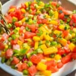A bowl of colorful salsa made with diced watermelon, mango, green bell pepper, red onion, and fresh herbs, sitting on a wooden table in natural sunlight.