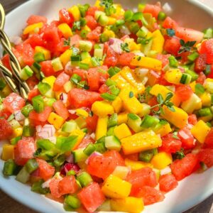 A bowl of colorful salsa made with diced watermelon, mango, green bell pepper, red onion, and fresh herbs, sitting on a wooden table in natural sunlight.