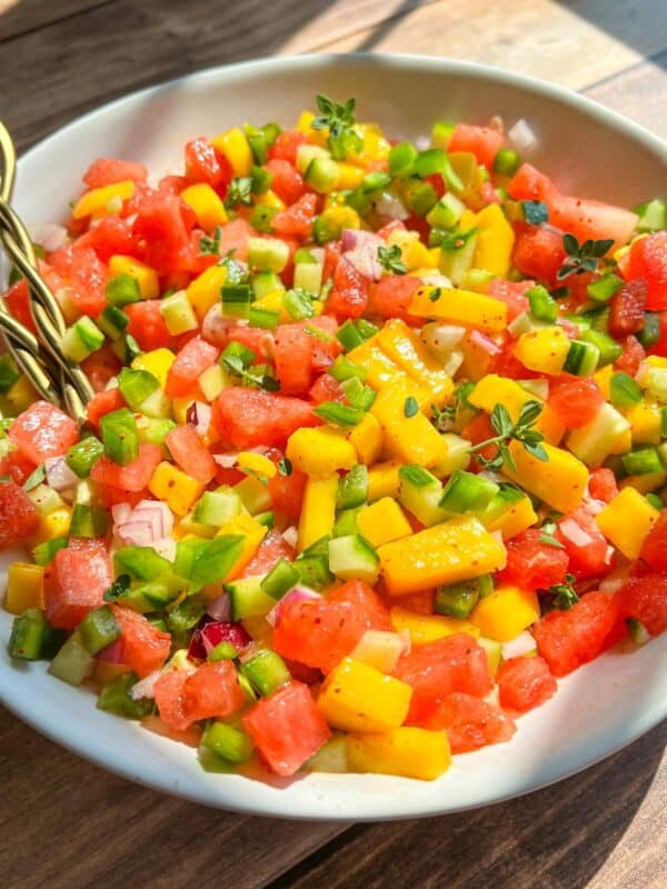 A bowl of colorful salsa made with diced watermelon, mango, green bell pepper, red onion, and fresh herbs, sitting on a wooden table in natural sunlight.