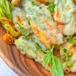 A wooden bowl filled with fried zucchini flowers, some garnished with fresh basil and herb sprigs. The blossoms are lightly battered and crispy, with hints of orange and green on the petals.