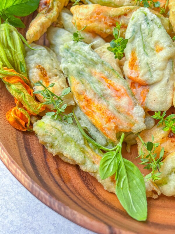 A wooden bowl filled with fried zucchini flowers, some garnished with fresh basil and herb sprigs. The blossoms are lightly battered and crispy, with hints of orange and green on the petals.