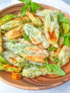 A wooden plate filled with golden, crispy fried zucchini blossoms, inspired by a Grilled Zucchini Recipe, garnished with fresh green herbs and basil leaves.