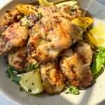 A bowl of herb marinated chicken pieces garnished with fresh herbs and lemon wedges, with visible grill marks and seasoning, bathed in natural sunlight.