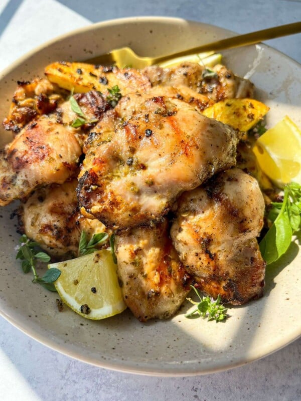 A bowl of herb marinated chicken pieces garnished with fresh herbs and lemon wedges, with visible grill marks and seasoning, bathed in natural sunlight.