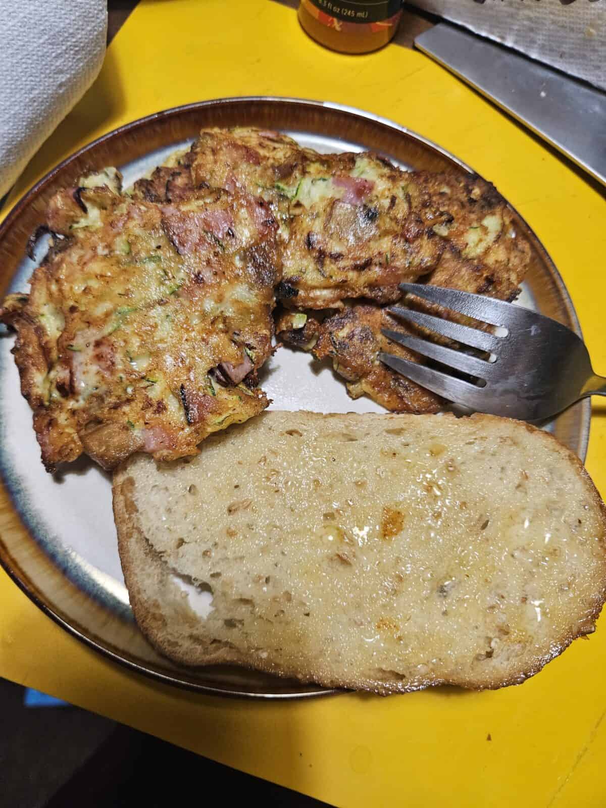 A plate with two vegetable fritters and a slice of bread with butter or spread. A fork rests on the plate. The background shows a yellow table surface and a paper towel.