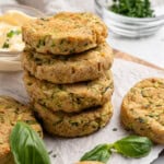 A stack of golden, freshly baked zucchini fritters on parchment paper, surrounded by fresh basil leaves. A small bowl of mayonnaise and a bowl of chopped herbs are in the background, with a whole zucchini partially visible.